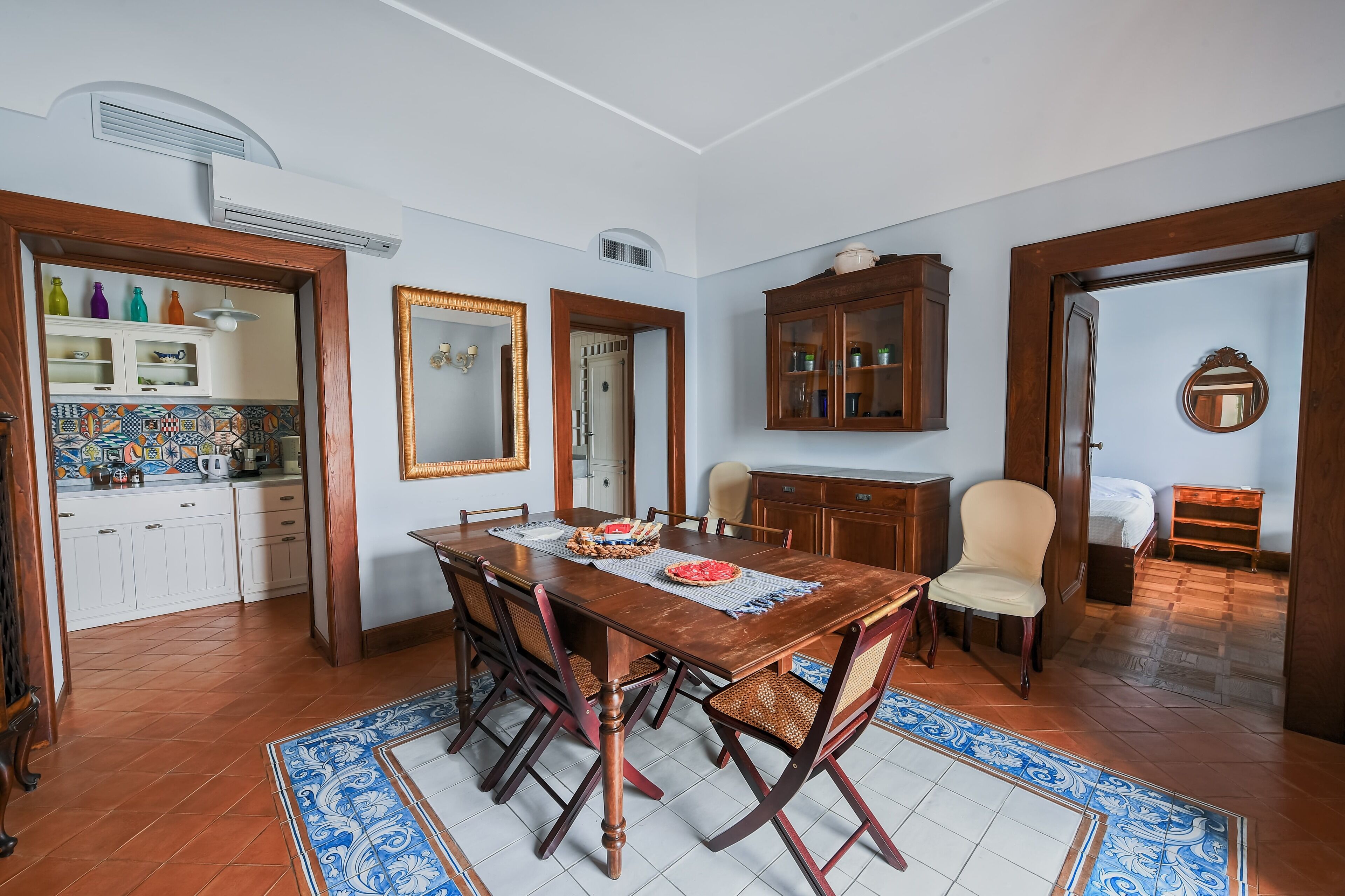 Un comedor con mesa de madera, sillas y piso de baldosas. Se observa una zona de cocina con armarios y salpicadero de azulejos.