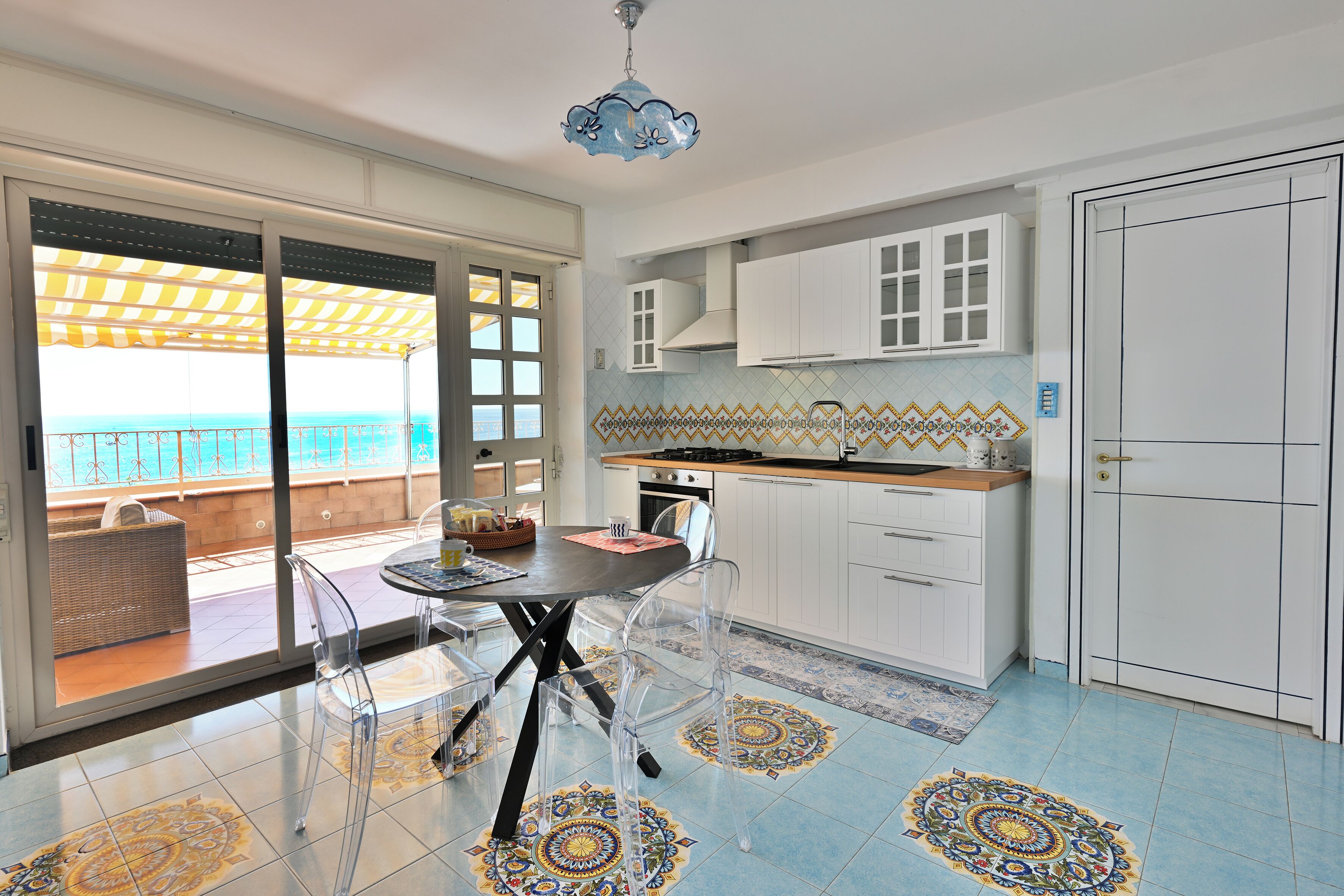Cozinha moderna com armários brancos, mesa de jantar com cadeiras transparentes e vista para o mar através de grandes portas de vidro deslizantes.