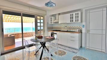 Cozinha moderna com armários brancos, mesa de jantar com cadeiras transparentes e vista para o mar através de grandes portas de vidro deslizantes.