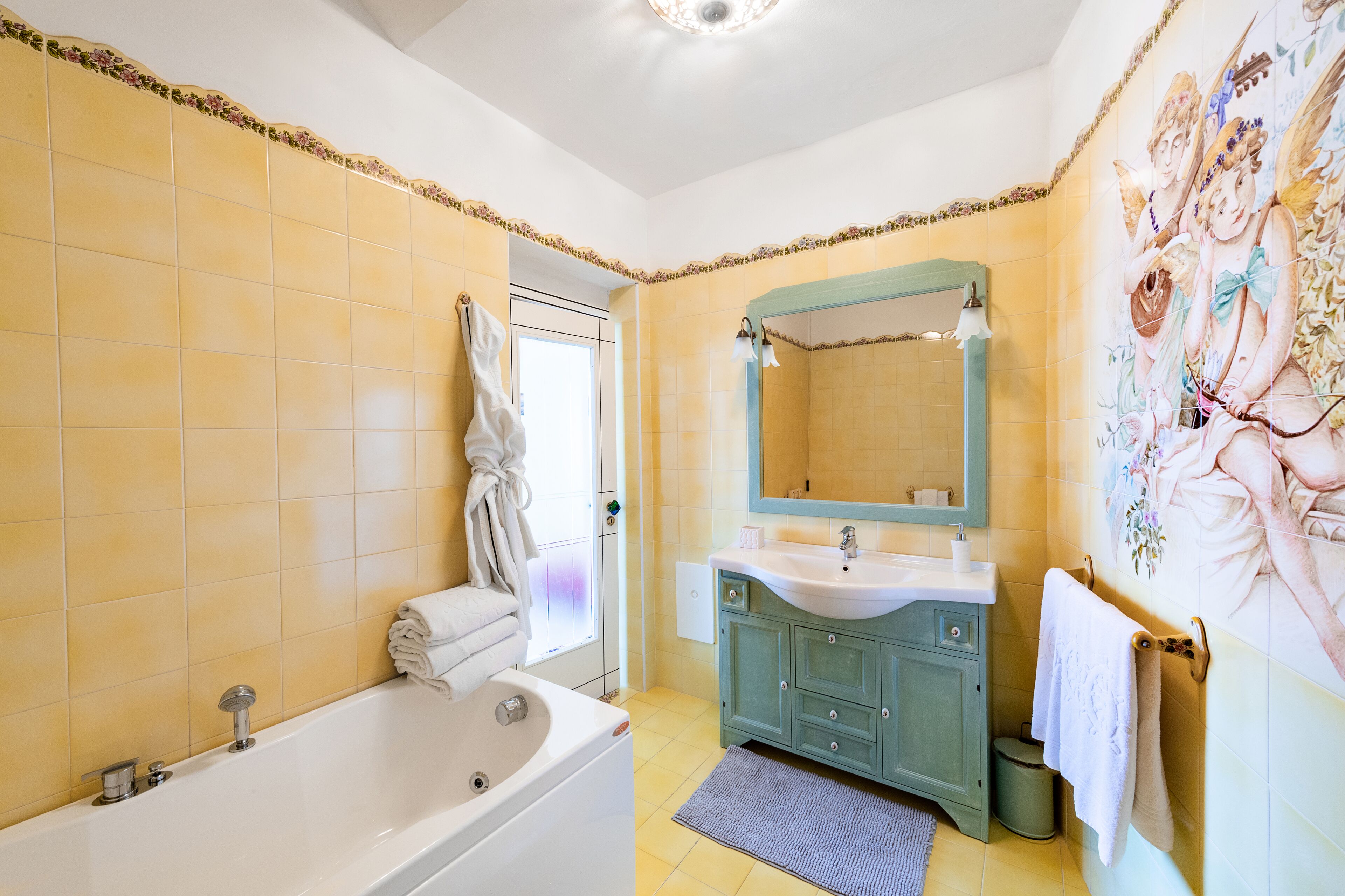 Ein Badezimmer mit gelben Fliesen, einer weißen Badewanne, einem Waschbecken mit blauem Unterschrank und einem Wandgemälde.
