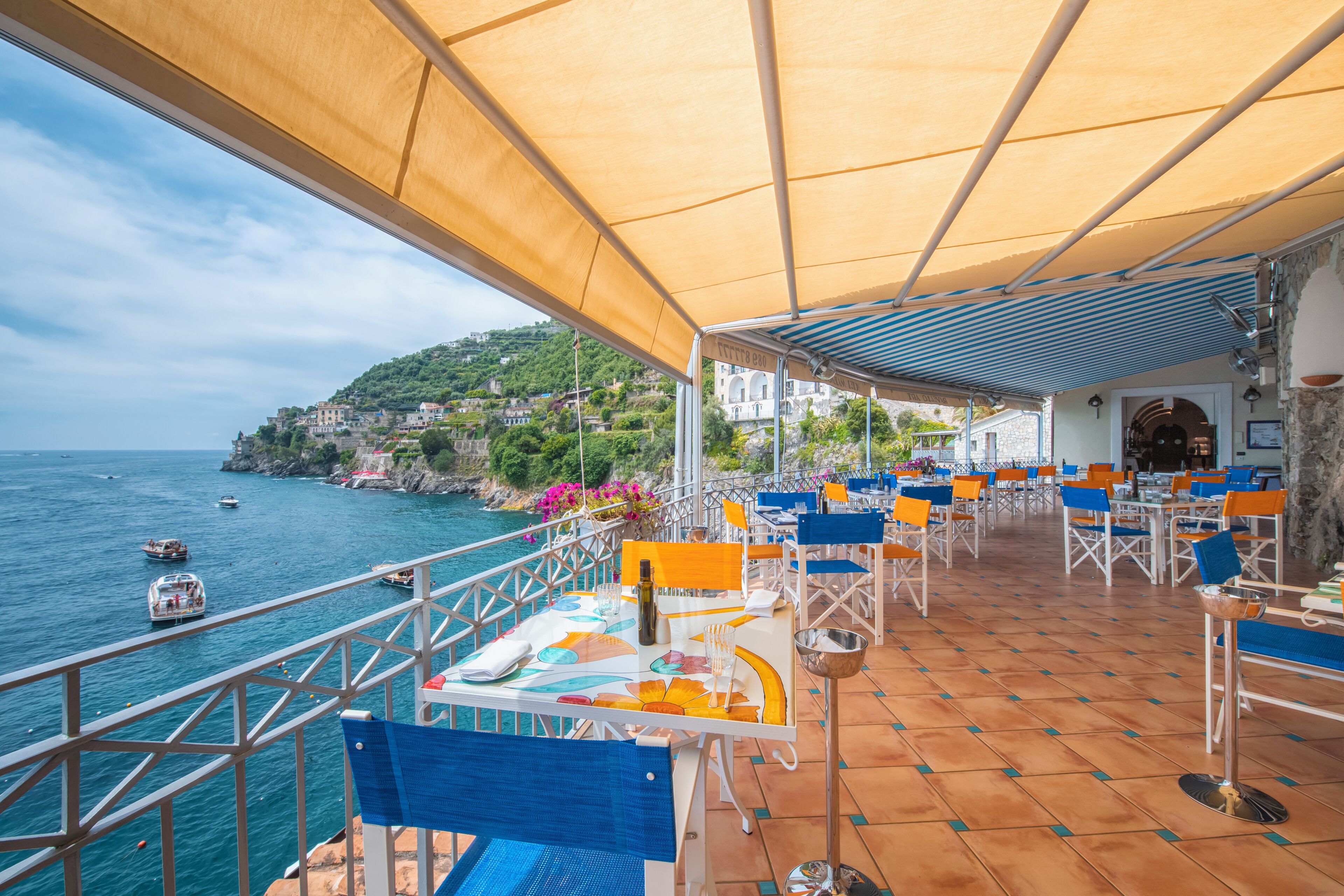 An outdoor terrace with tables and chairs, overlooking a coastal area with boats and buildings.