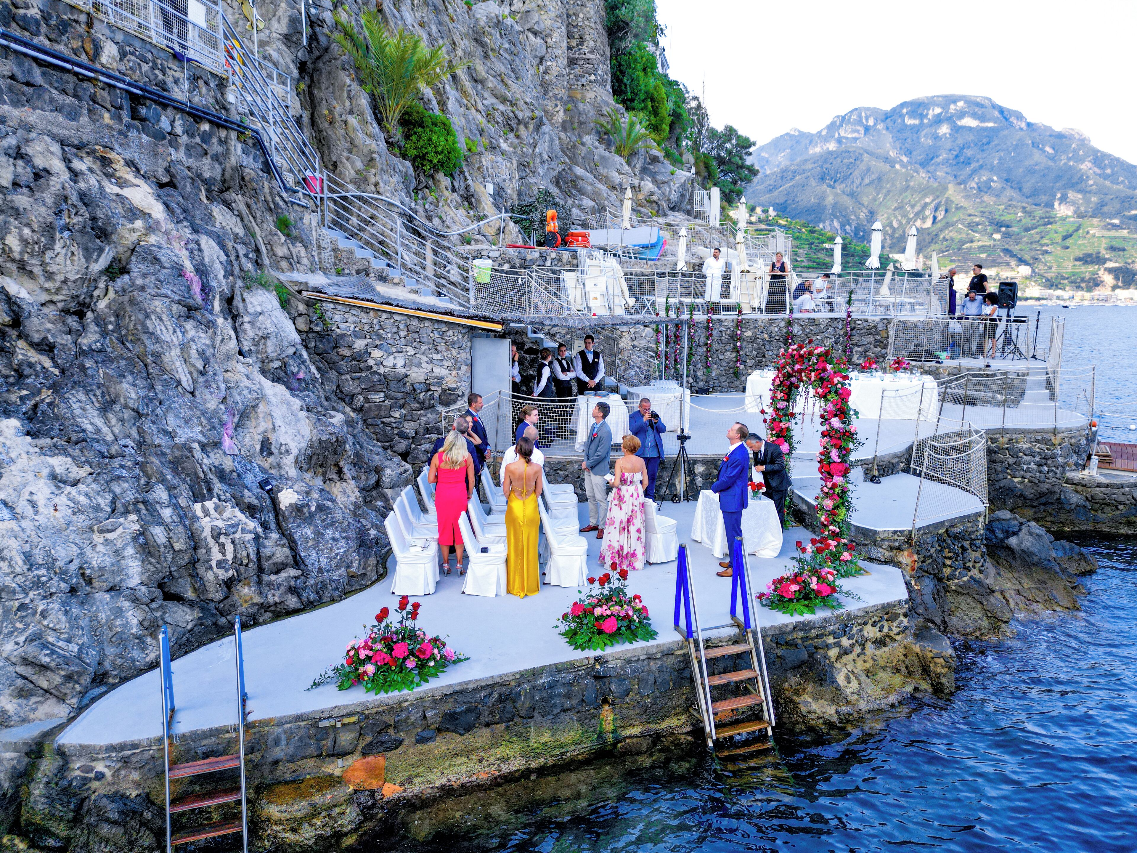 A wedding ceremony on a rocky waterfront with a stage, guests, and a mountainous backdrop.