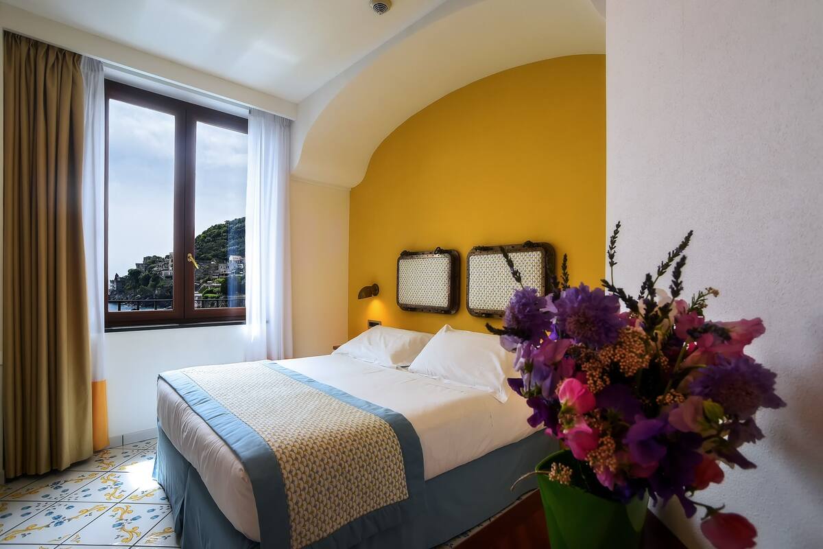 Une chambre d’hôtel avec un lit, une fenêtre avec des rideaux, un mur jaune et un arrangement floral.