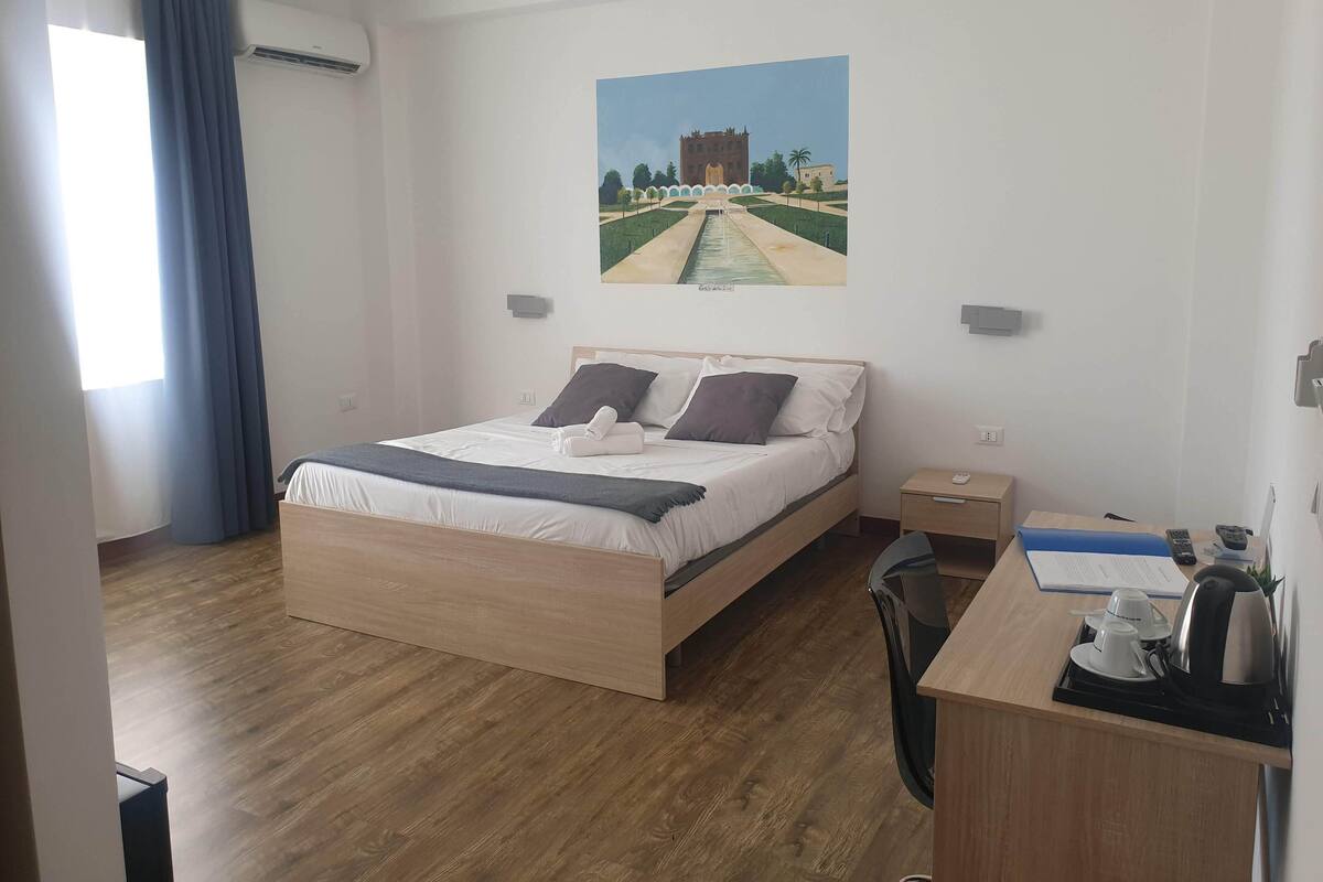 Hotellihuone, jossa on sänky, työpöytä, jossa on vedenkeitin, kuppeja ja kaukosäädin.
