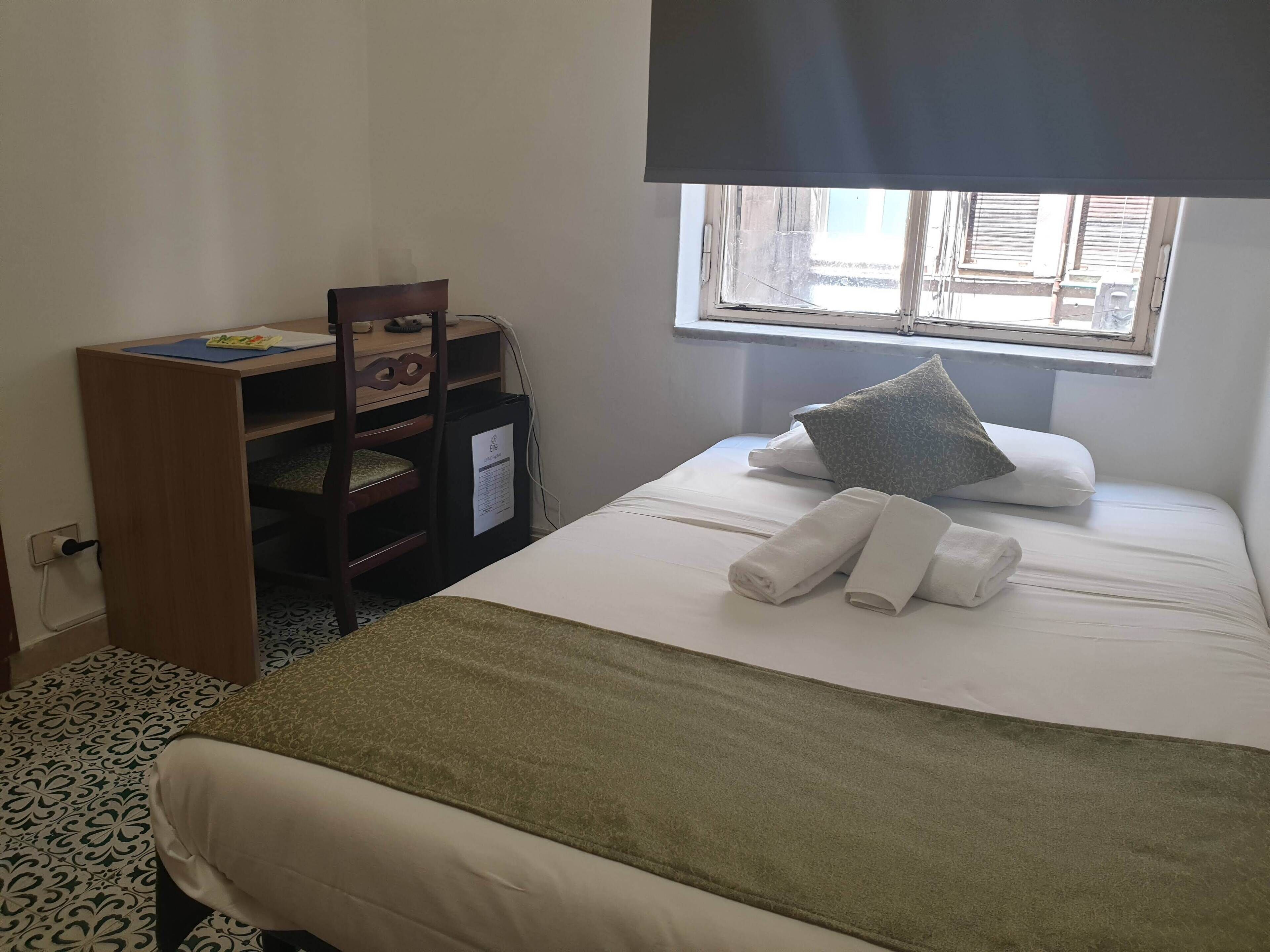 A neatly made bed with towels, a desk with a chair, and a window with blinds.