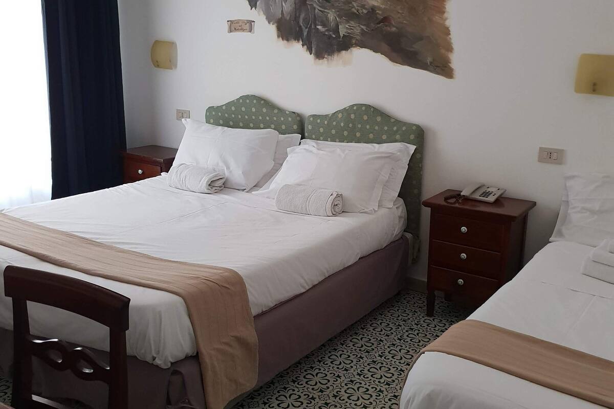 Hotellihuone, jossa on suuri sänky, tuoli, pieni pöytä ja koristeellinen seinämaalaus.