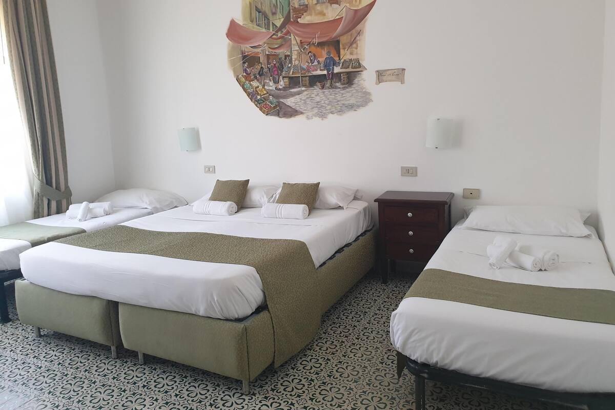Hotellihuone, jossa on kolme sänkyä, kuviollinen lattia, seinämaalaus ja pieni puinen yöpöytä.