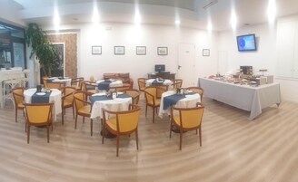 Zona pranzo con tavoli e sedie rotonde, un tavolo per il buffet con cibo e una televisione a parete.