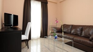A modern living room with a brown leather sofa, a glass coffee table, a white chair, a TV on a black stand, and a window with brown curtains. - E H SUITES ROME AIRPORT (Fiumicino)