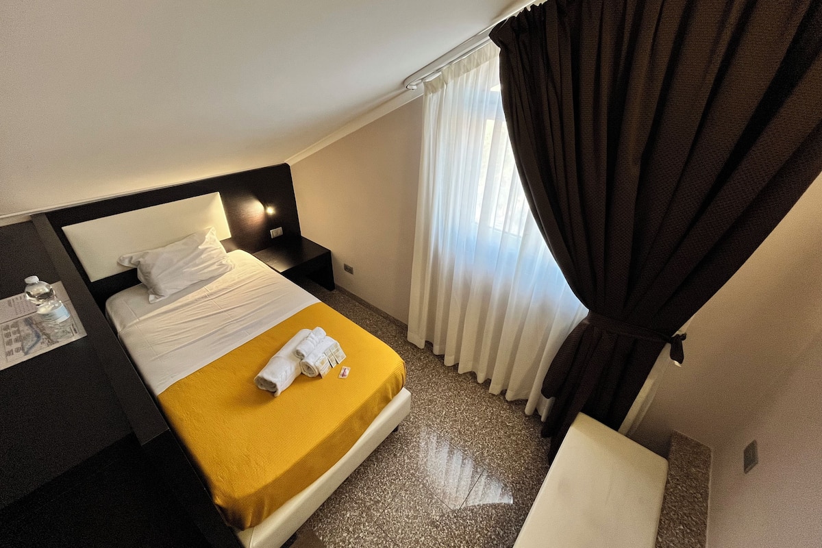 A hotel room with a bed, a nightstand, a desk with a lamp, a window with curtains, and a small table.