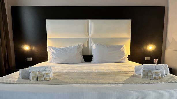 A neatly made bed with pillows, a folded towel, and a small card on the bed. - E H SUITES ROME AIRPORT (Fiumicino)