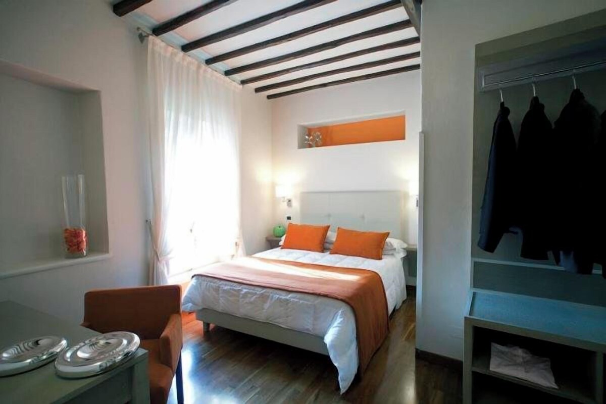 Una camera da letto con un letto, una sedia, una scrivania con piatti, un appendiabiti e una finestra con tende.