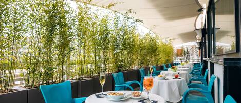 An outdoor dining area with white tables and blue chairs, surrounded by bamboo plants.