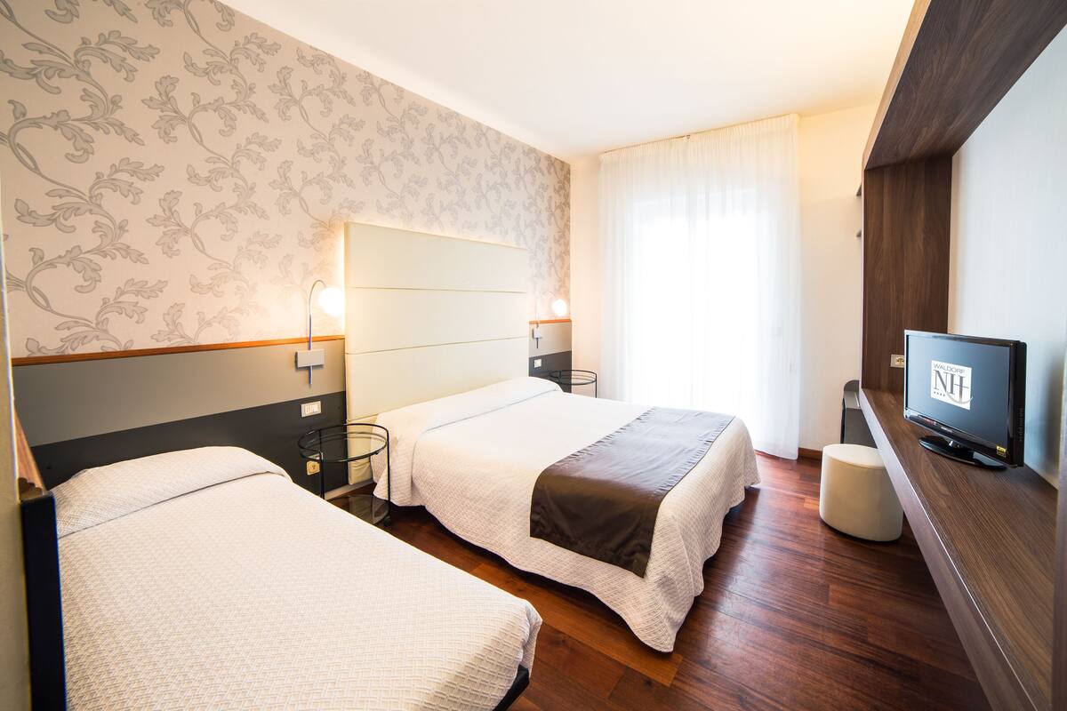 Ein Hotelzimmer mit zwei Betten, einem Fernseher, einem Schreibtisch und einem Fenster mit Vorhängen.