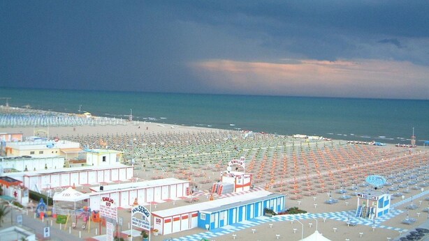 A beach with rows of umbrellas and beach chairs, a lifeguard tower, and buildings along the shore. - Hotel Artide (Rimini)