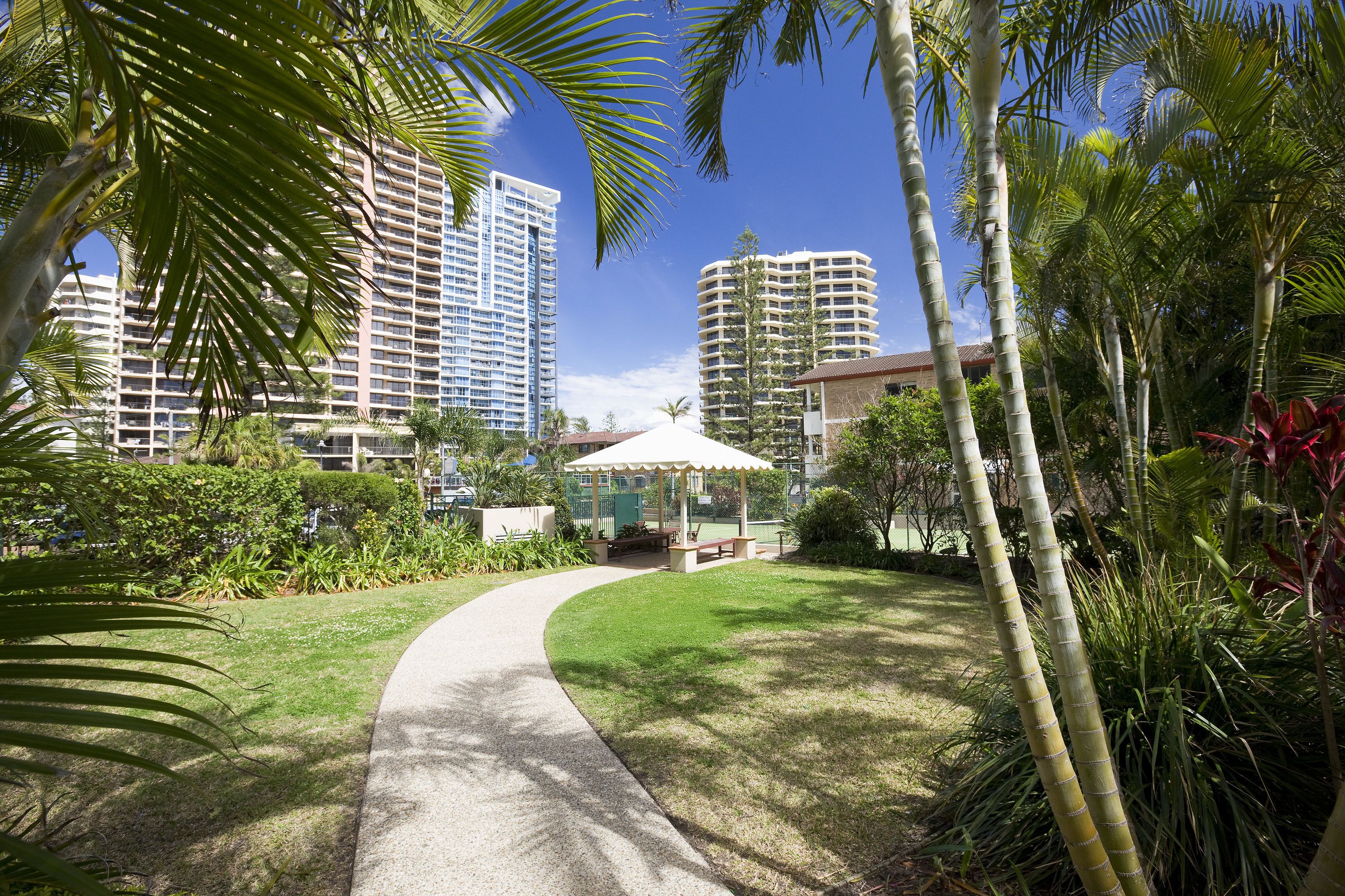 A pathway leading to a gazebo surrounded by lush greenery and tall buildings in the background.