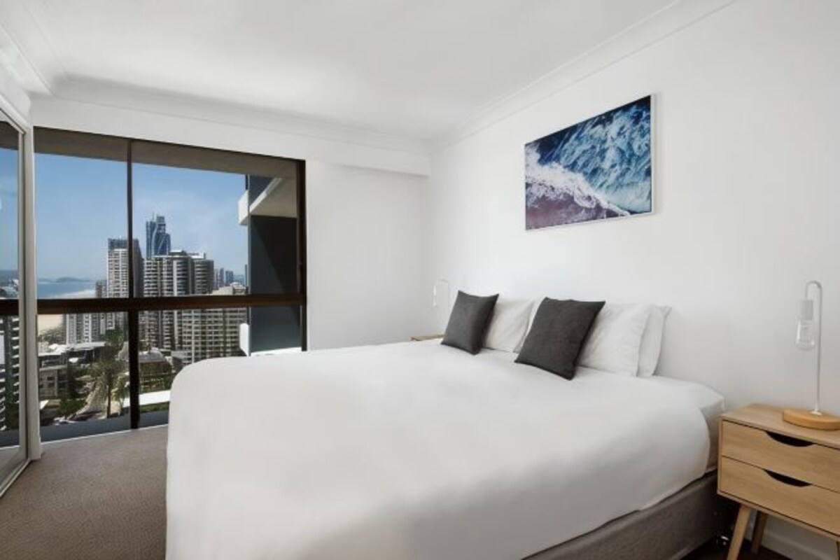 A hotel room with a large bed, a nightstand, a window with a city view, and a painting on the wall.