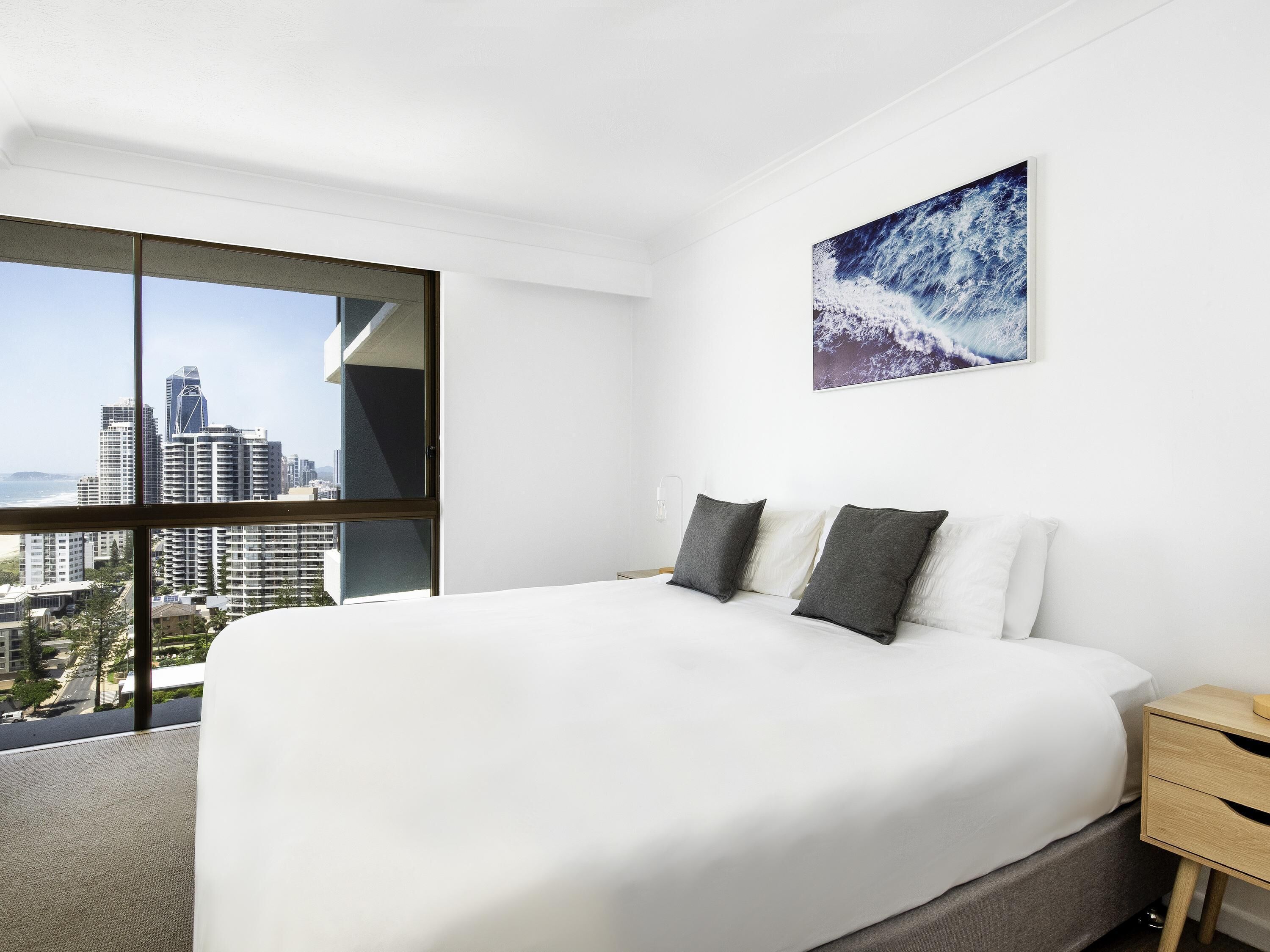 A hotel room with a large bed, a nightstand, a wall-mounted painting, and a city view through a large window.