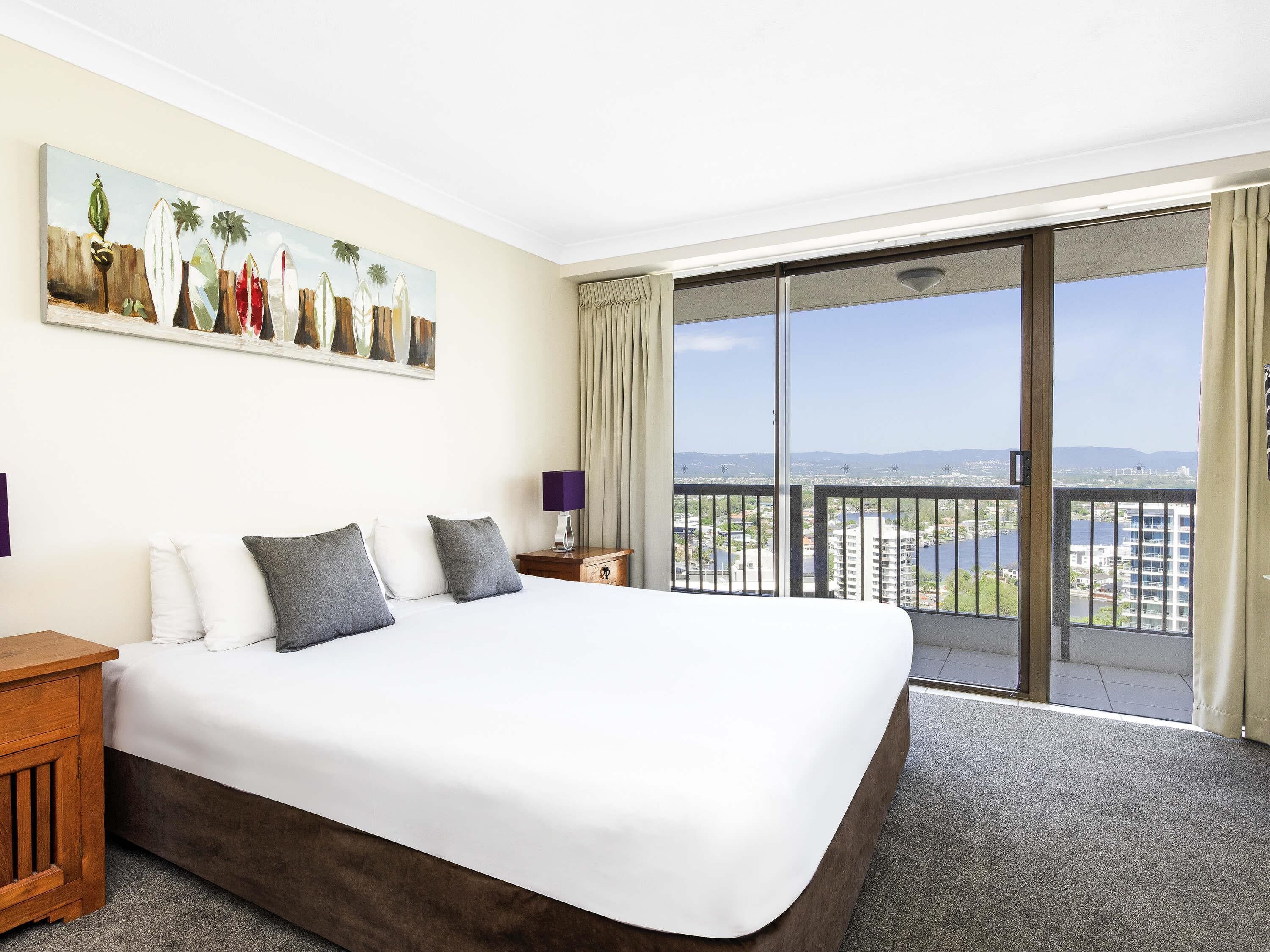 Ein Hotelzimmer mit einem großen Bett, einem Nachttisch, einem Gemälde und einem Balkon mit Blick auf die Stadt.
