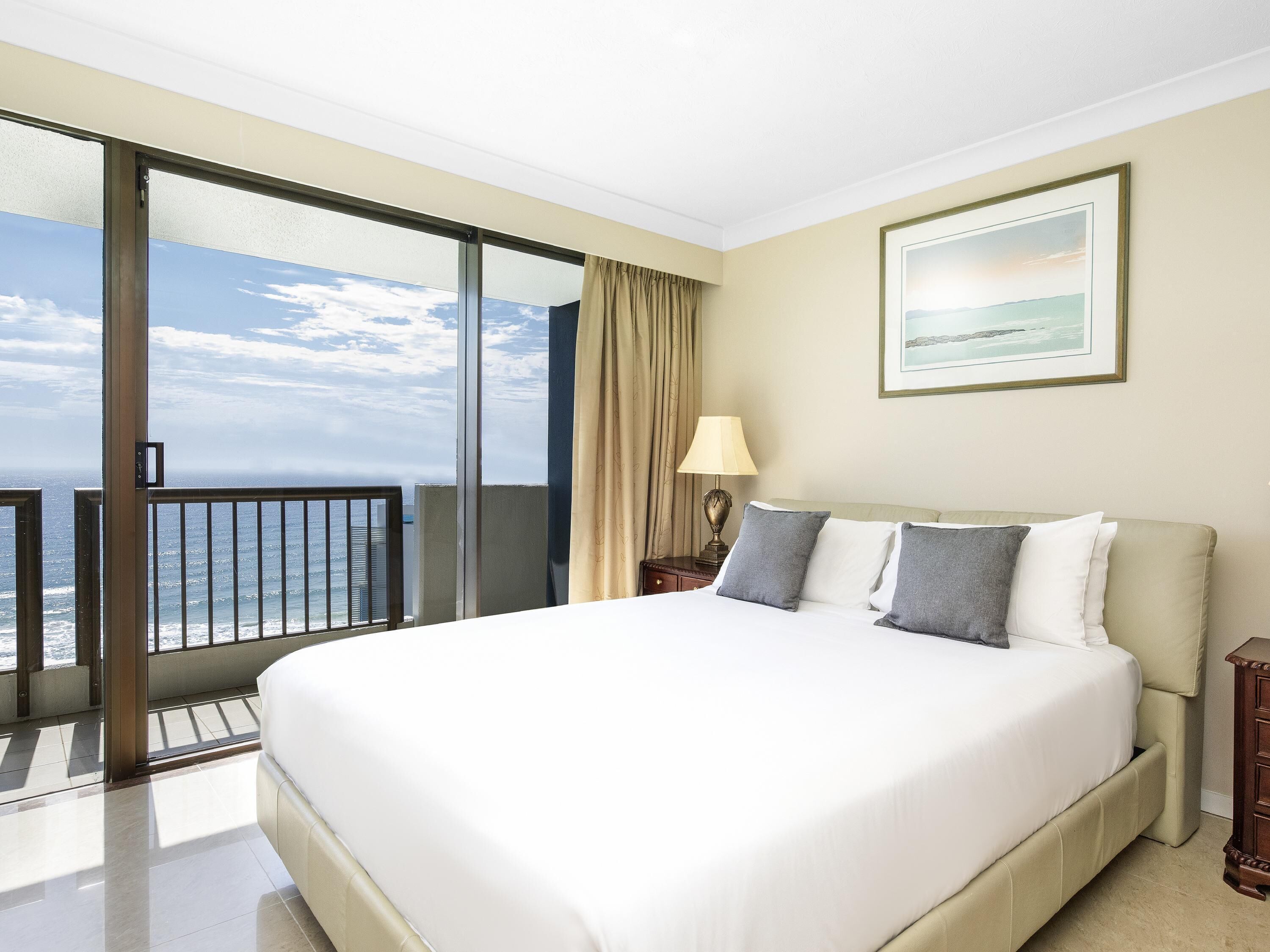 A hotel room with a large bed, a nightstand, a lamp, a framed picture, and a balcony with a view of the ocean.