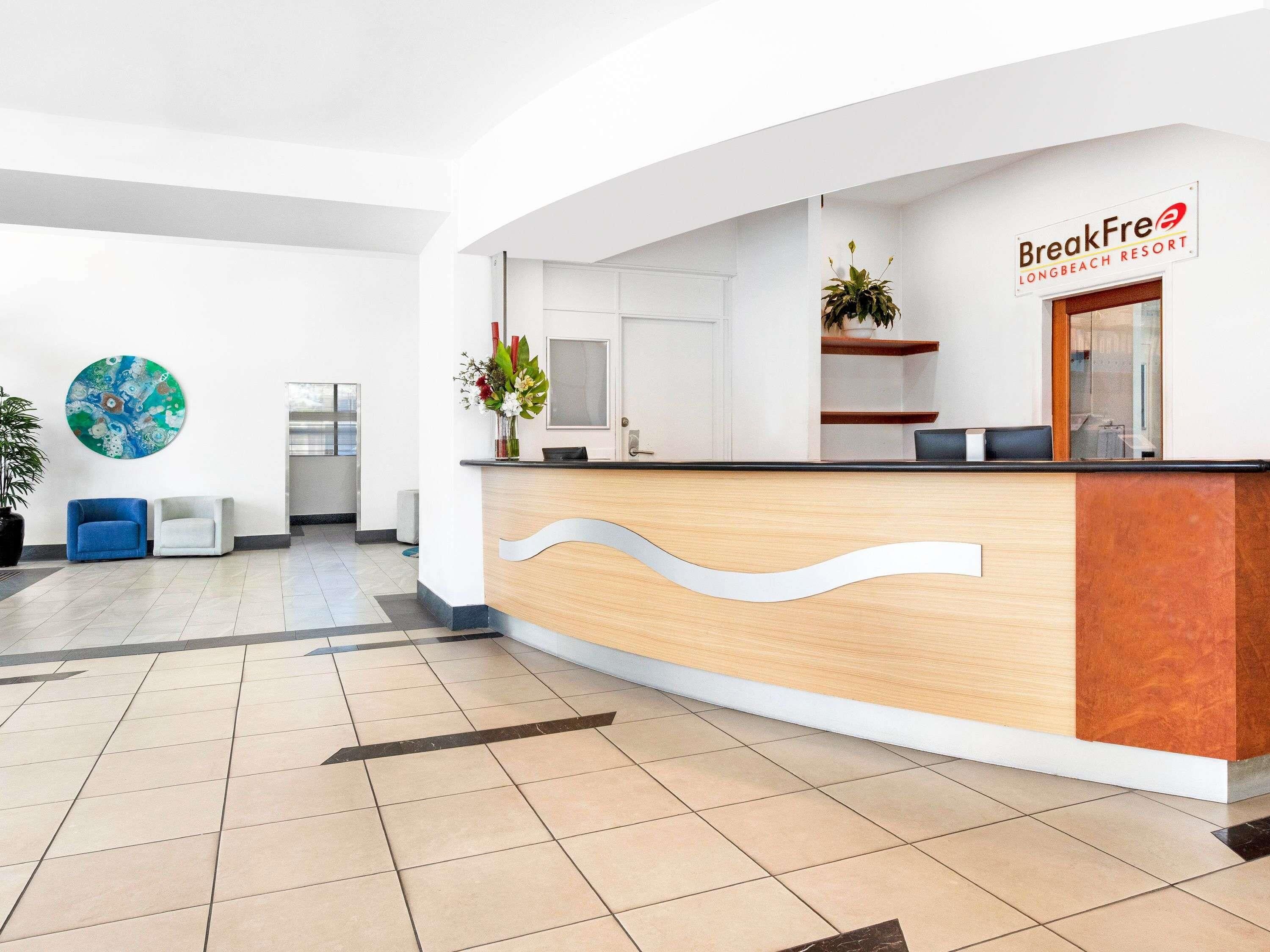 A reception area with a wooden reception desk, a blue chair, a white chair, a large abstract painting, and a potted plant.