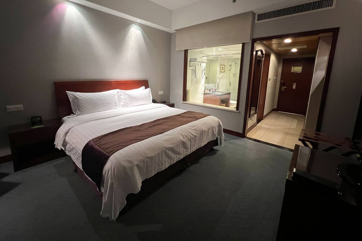 Habitación de hotel con una cama grande, una mesita de noche, una ventana que da a un baño y una puerta que conduce a otra habitación.