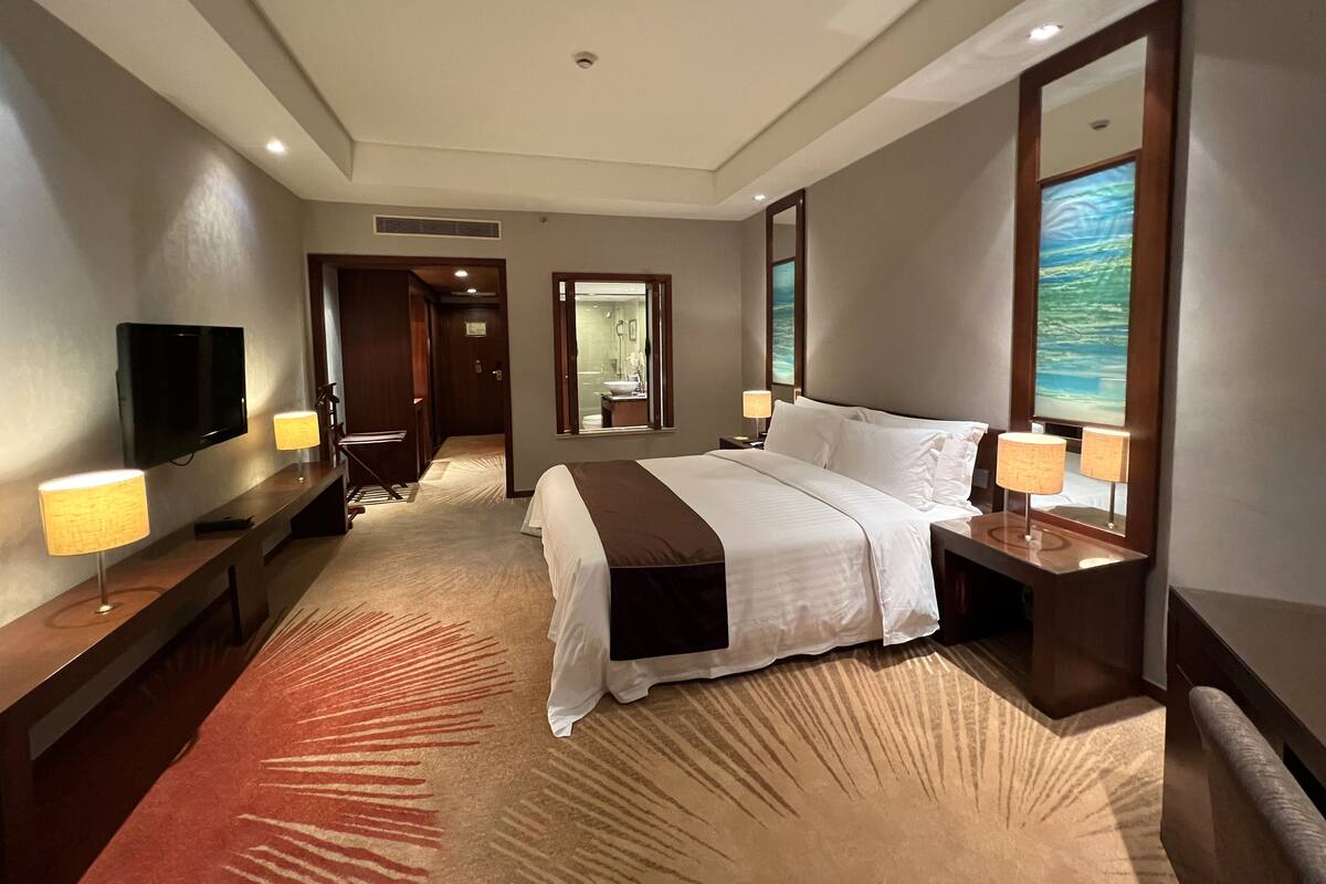 Habitación de hotel con cama, lámparas de noche, televisor, espejo de baño y vista al océano.