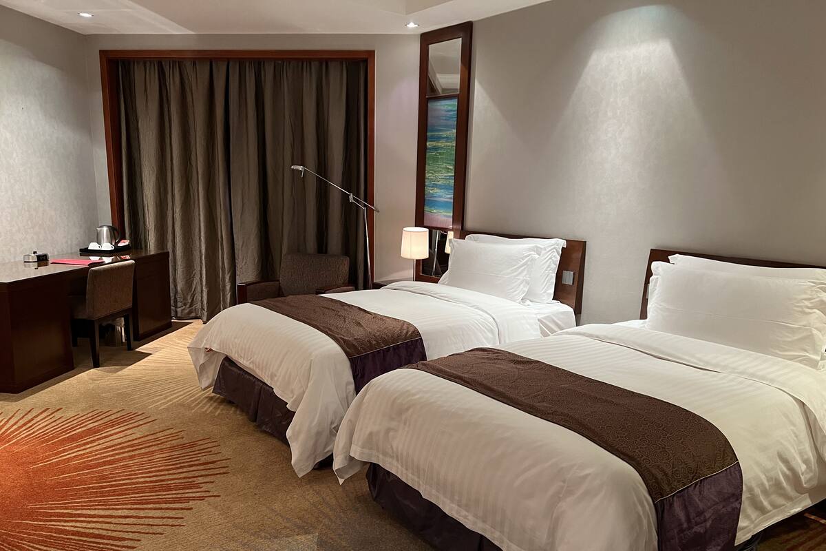 Habitación de hotel con dos camas, un escritorio, una silla, una lámpara y un ventanal grande con cortinas.