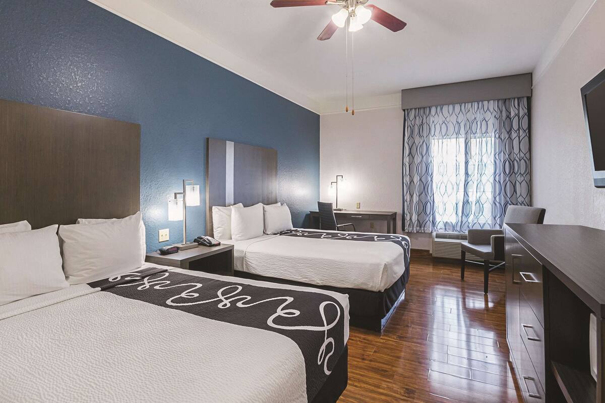A hotel room with two beds, a desk, a chair, a ceiling fan, and a large window with curtains.