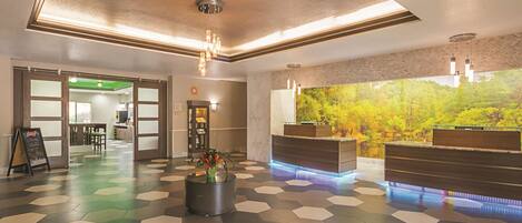 A hotel lobby with a reception desk, a large window with a forest view, a patterned floor, and a green-lit reception area.