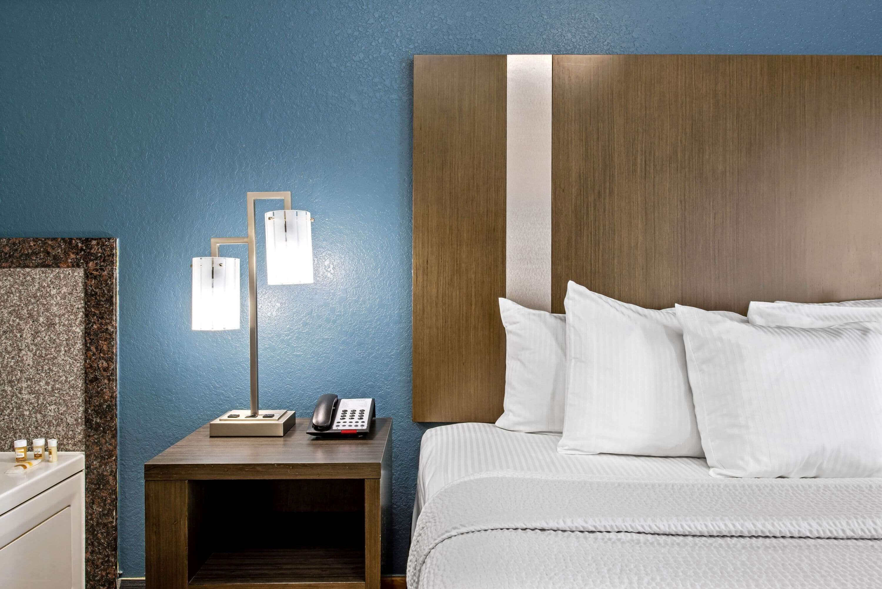 A hotel room with a bed, a nightstand with a lamp, a phone, and a small table with bottles.