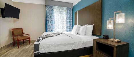 Habitación de hotel con una cama, una silla, un televisor montado en la pared y una mesita de noche con una lámpara.