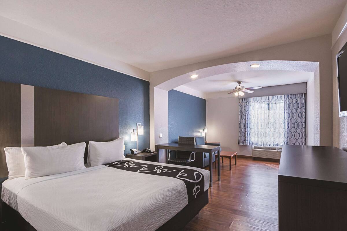 A hotel room with a bed, a desk, a chair, a ceiling fan, and a window with curtains.