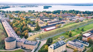 Eine Vogelperspektive zeigt einen historischen Gebäudekomplex mit mehreren Strukturen, darunter ein großer Turm und einige kleinere Gebäude, umgeben von offenen Feldern und dichter Baumlandschaft.