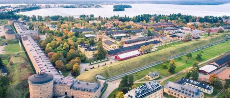 An aerial view of a historic building complex with multiple structures, including a large tower and several smaller buildings, surrounded by a mix of open fields and dense trees.
