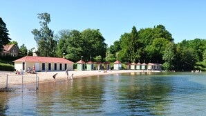 Ein Seeufer mit kleinen Gebäuden, einem Sandstrand und Menschen, die sich an Wassersportaktivitäten beteiligen.