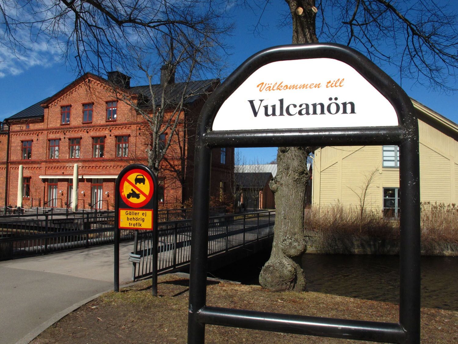 A sign with 'Vulkanön' and 'Välkommen till' indicating a welcoming area, a no parking sign, and a building with a red brick facade.