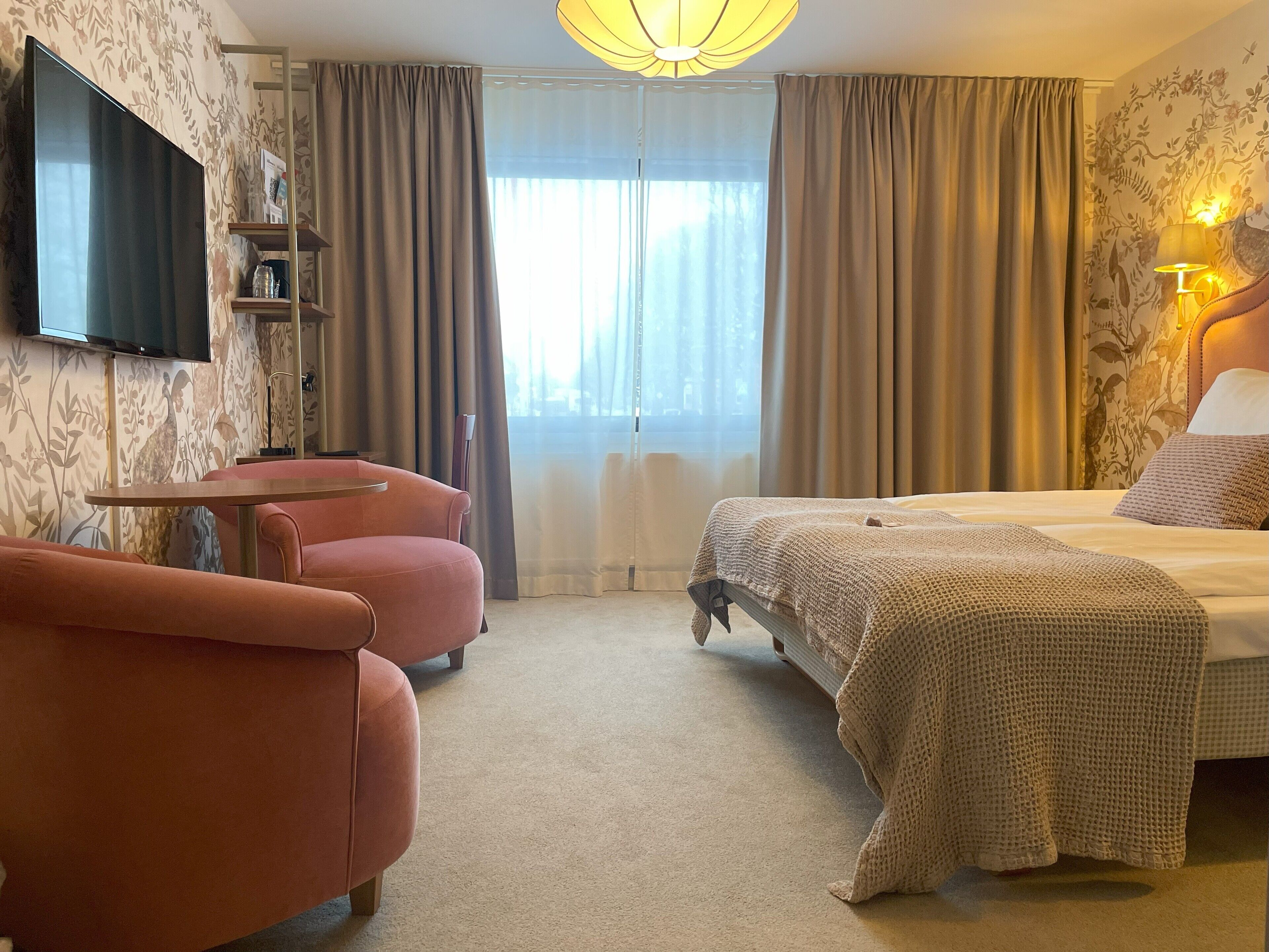 A hotel room with a bed, a pink armchair, a small table, a TV mounted on the wall, and a window with curtains.