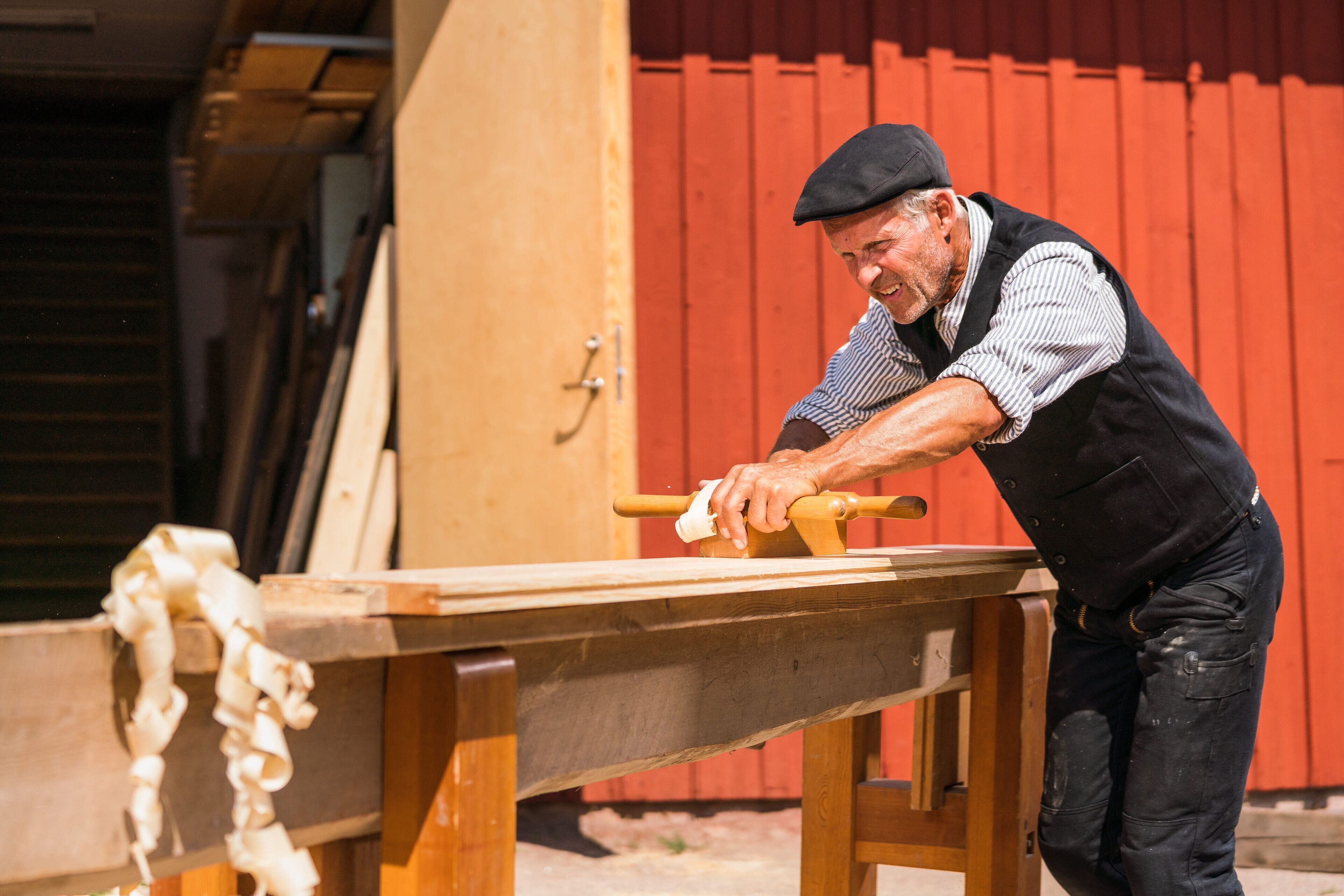 Une personne utilise un outil en bois sur une table à l’extérieur, avec un bâtiment rouge en arrière-plan.