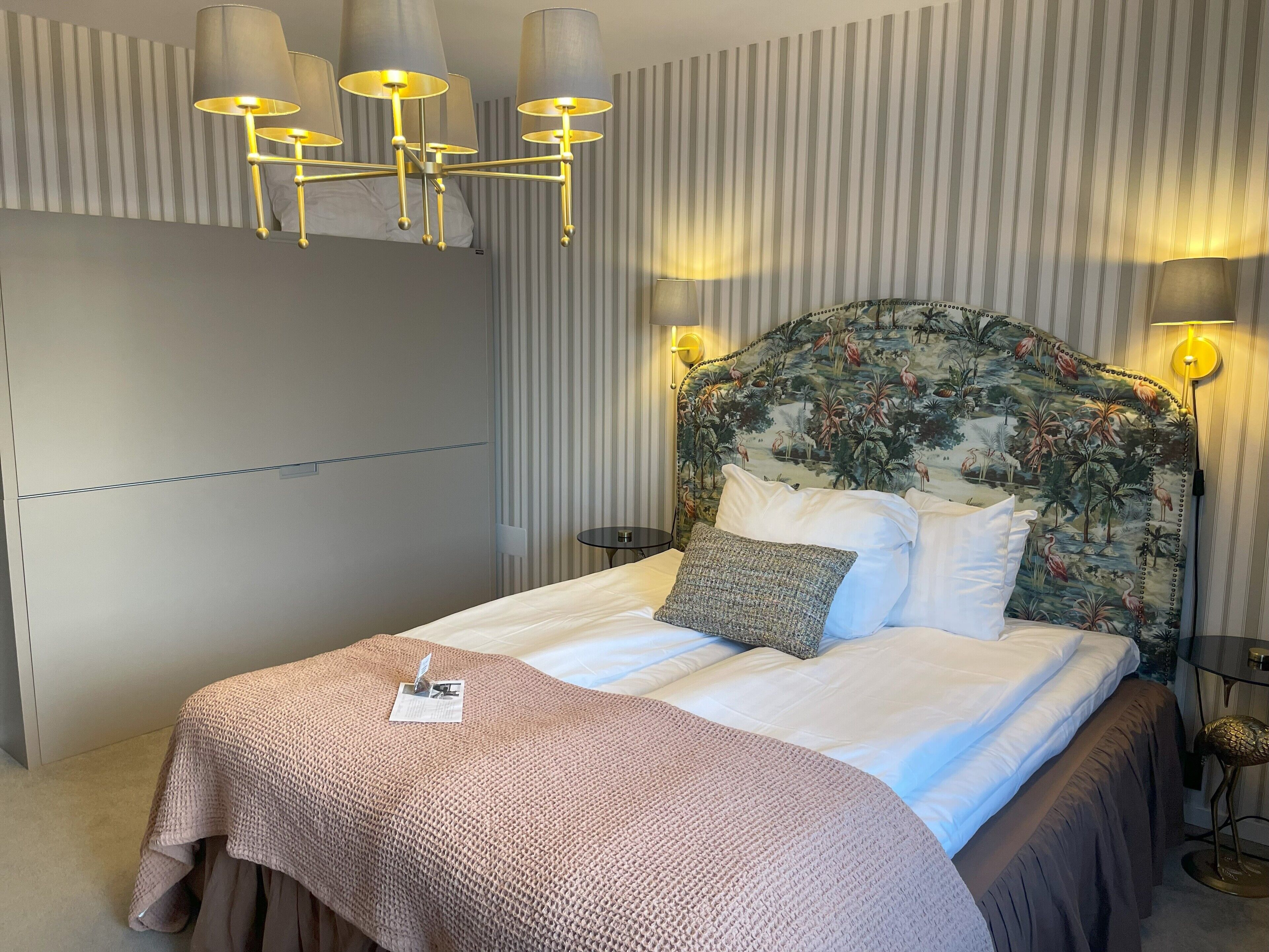 Una cama bien hecha con un cabecero floral, una mesita de noche y una lámpara de pared.