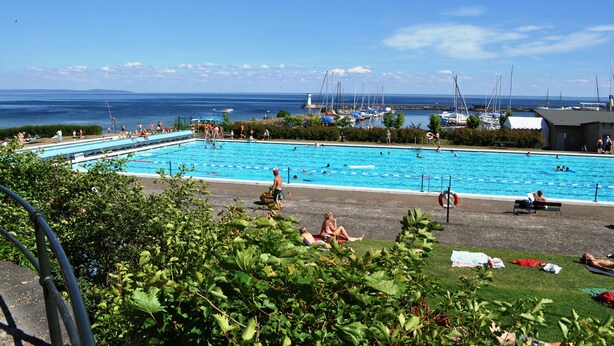 Ein Schwimmbecken mit darin schwimmenden und sonnenden Personen, umgeben von viel Grün und Blick aufs Meer.