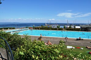 Una piscina con gente nadando y tomando el sol, rodeada de vegetación y vistas al mar.