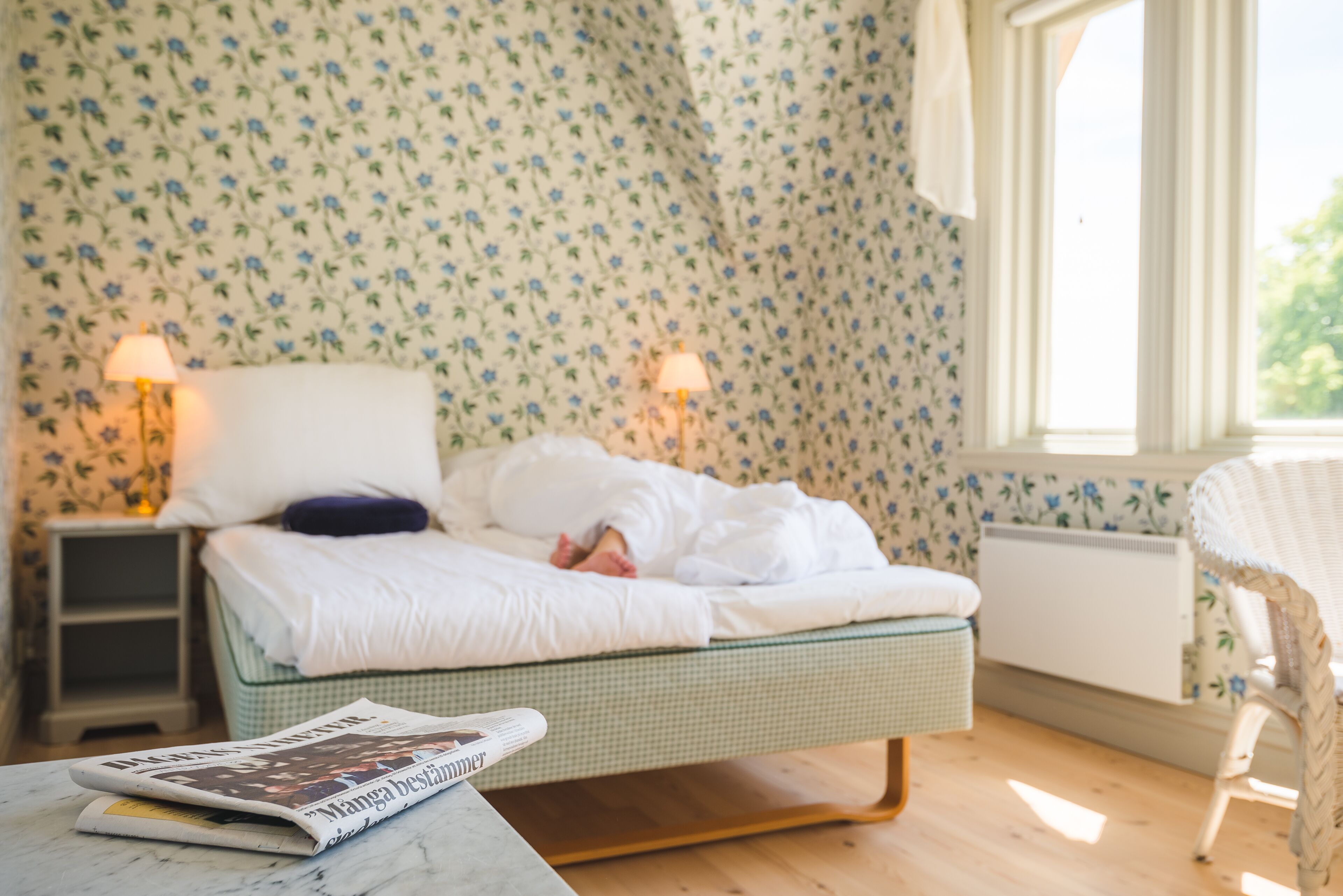 Un dormitorio con una cama, dos lámparas de noche, una silla y una mesa con una revista.