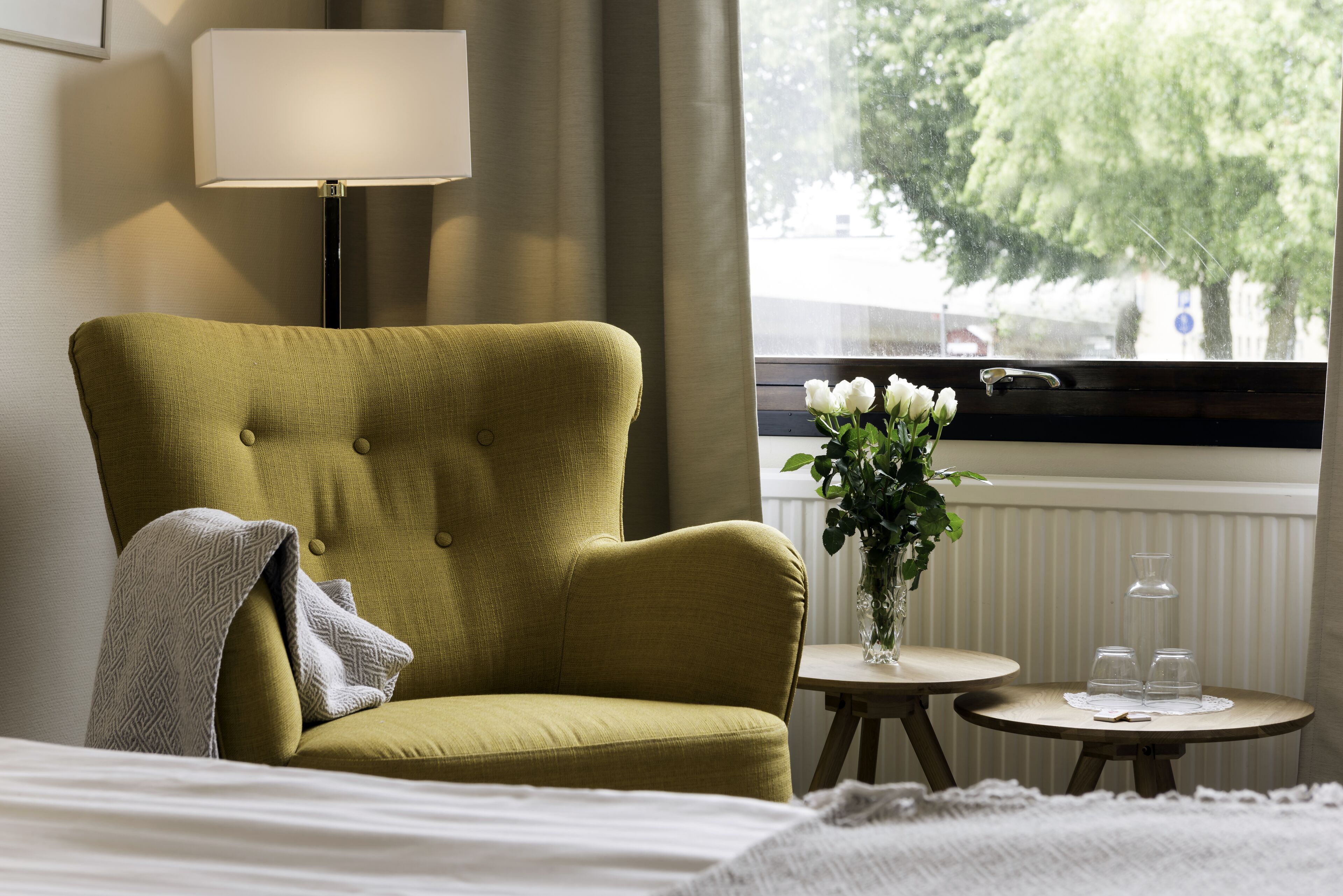 A hotel room with a green armchair, a bedside table with a vase of flowers, and a window with a view of trees.