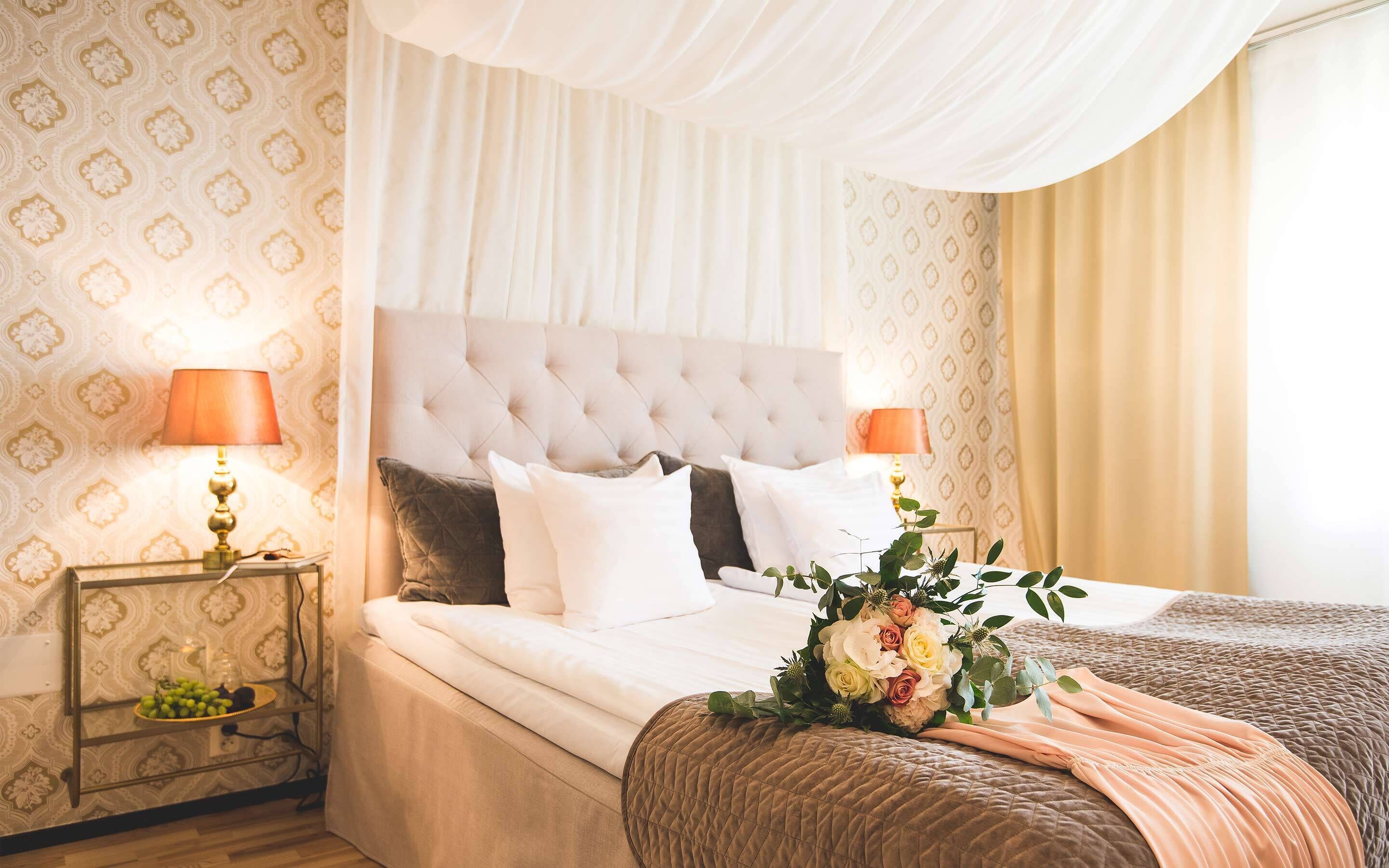 Een slaapkamer met een gestapeld hoofdbord, wit beddengoed, twee nachtkastjes met lampen en een bloemstuk op het bed.