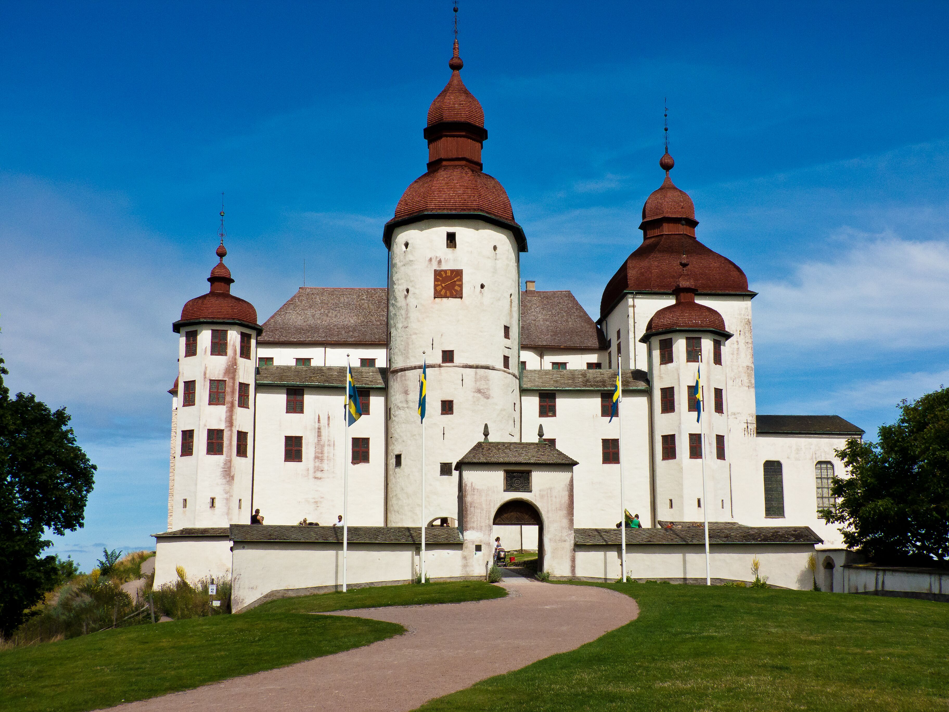 Ein historisches Gebäude mit einem zentralen Turm und zwei kleineren Türmen, einem gepflasterten Eingangsbereich und einer umlaufenden Grünfläche.