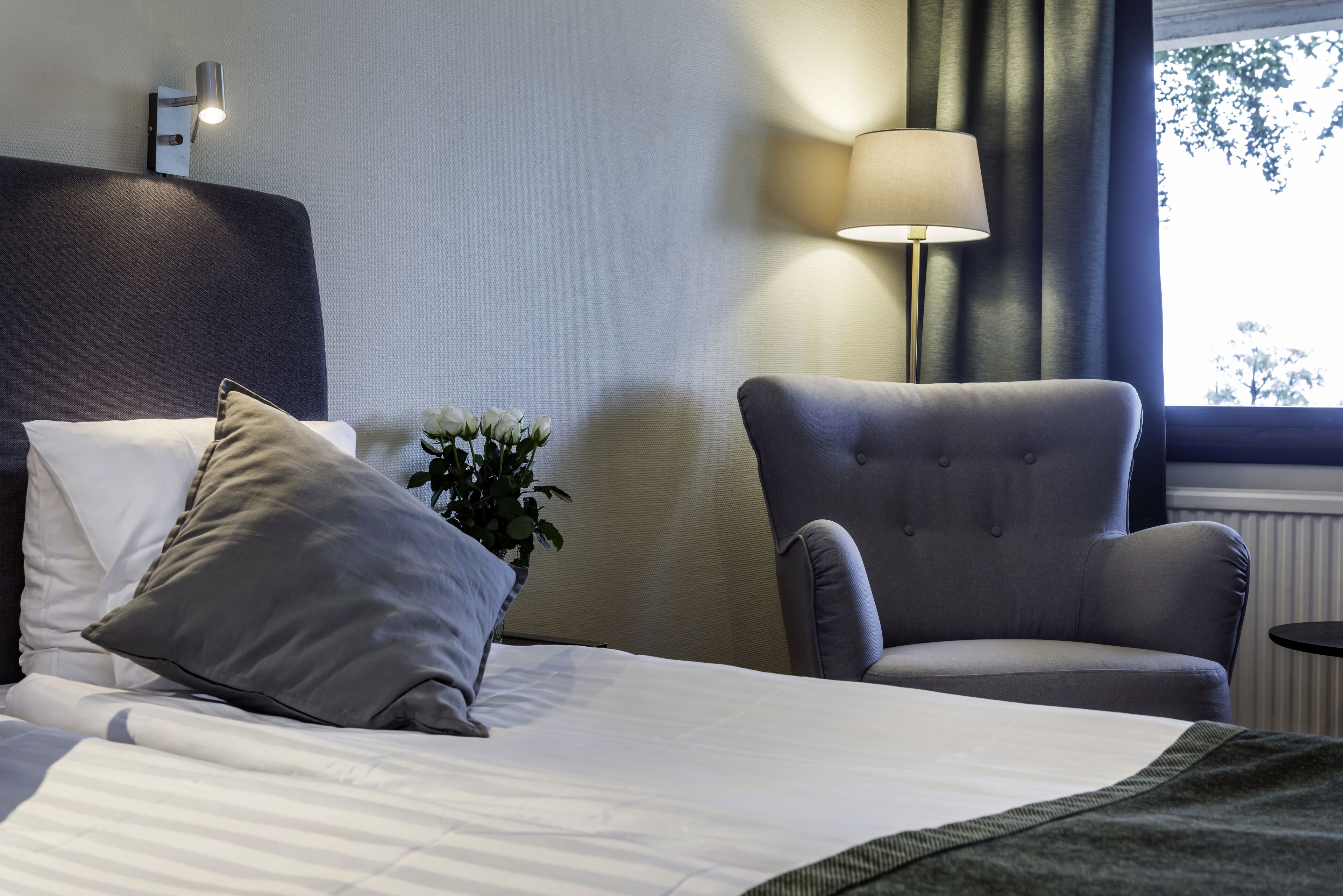 Una habitación de hotel con una cama, un sillón gris, una lámpara y una ventana con cortinas.