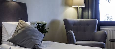 Una habitación de hotel con una cama, un sillón gris, una lámpara y una ventana con cortinas.