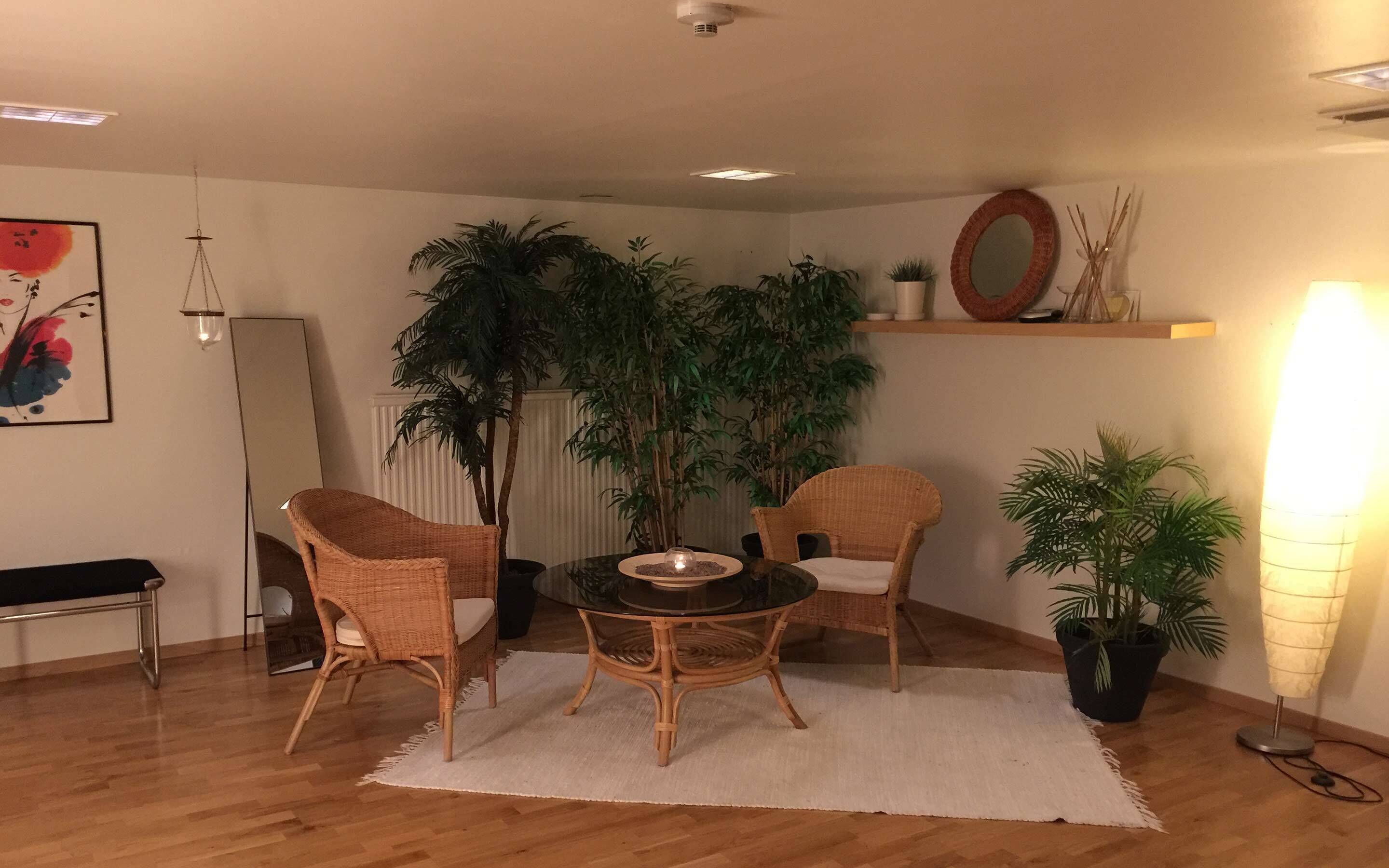 A living room with a round table, two wicker chairs, a white rug, and several potted plants.