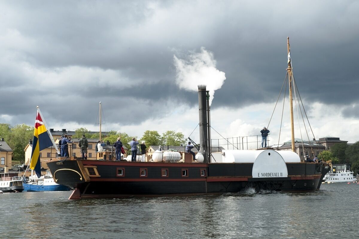 A steamboat named E. Norderval II docked at a harbor with a flag, people on board, and a cloudy sky in the background.